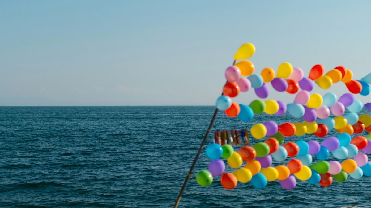 A bunch of brightly coloured balloons tied above a rocky shoreline beside the ocean.