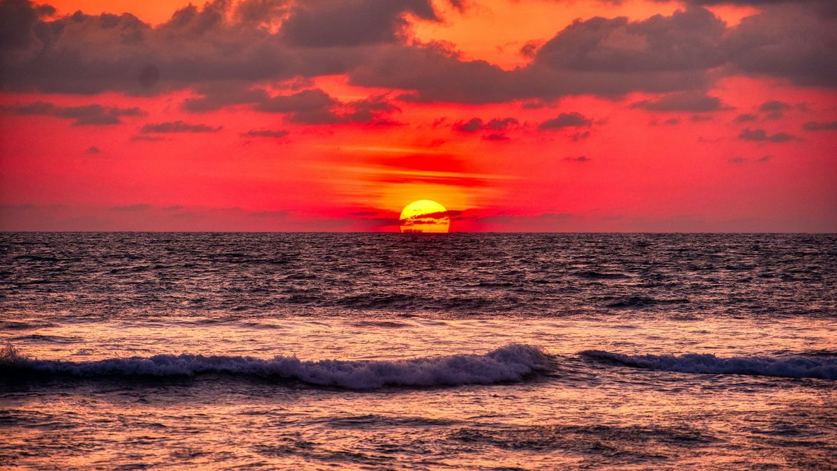 A deep orange sunset over the ocean, with waves reaching a dark shoreline.