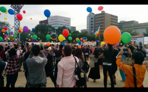 A crowd at an outdoor ceremony watching dozens of coloured balloons rise into the sky.