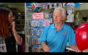 Carly Wilson interviewing a balloon retailer in his shop, surrounded by foil balloons and party supplies.